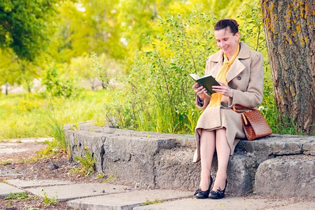 围巾女摄影照片_优雅的女商人坐在石墙上在一个阳光明媚的春天，和阅读一本有趣的书