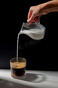 brew摄影照片_咖啡因和饮料概念--用水壶将奶油或牛奶倒到桌子上的一杯咖啡中。手拿着罐子把奶油倒到一杯咖啡里