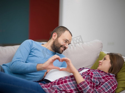 示爱男人摄影照片_快乐的男人和孕妇显示心脏标志与手指同时在家里的沙发上放松。男子和孕妇显示心脏征