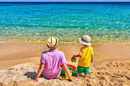 坐在爸爸的背上摄影照片_两岁的学步男孩和父亲在海滩上。充气戒指和沙滩玩具。暑假全家度假。希腊西索尼亚。