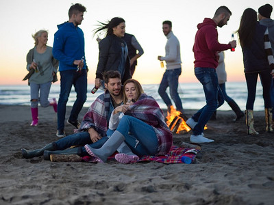 日落时分，一对年轻夫妇和朋友坐在海滩上围着篝火喝啤酒。一对情侣和朋友在海滩上享受日落