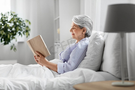 技术、老年和人的概念—高级妇女在家里卧室的床上看书。高级妇女阅读书在床上在家庭卧室