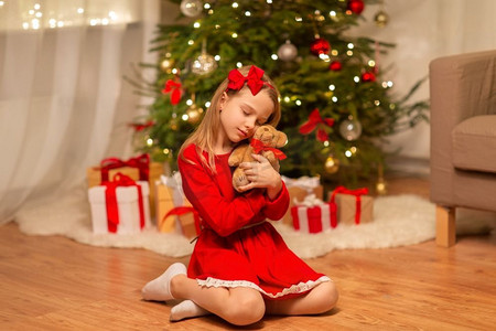 冬天穿的红摄影照片_圣诞节、节日和童年的概念--穿着红色连衣裙的女孩在家里拥抱着泰迪熊。穿红衣服的女孩在家里拥抱泰迪熊