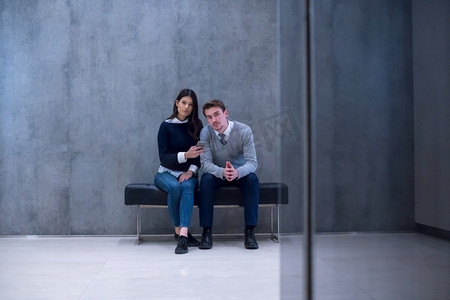 年轻的商务夫妇在休息时坐在办公室大堂的长椅上使用智能手机