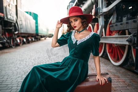 女人在红帽子坐在行李箱反对老式蒸汽火车。旧火车头。铁路发动机，古代铁路车辆