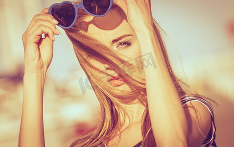 夏日时尚。戴着蓝色心形太阳镜的肖像女孩在码头的夕阳下享受夏日的微风