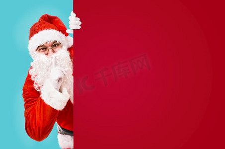 服装要求摄影照片_圣诞老人用手指放在嘴唇上，要求安静，上面有五颜六色的广告牌和文案空间。圣诞老人要求保持沉默