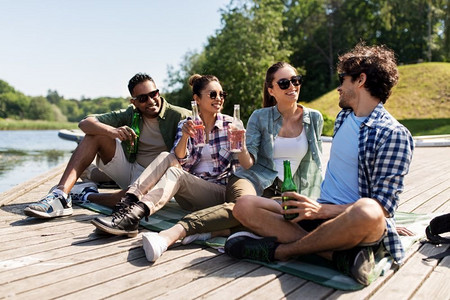 苹果酒摄影照片_休闲，野餐和人的概念—朋友喝啤酒和苹果酒在湖码头在夏季公园。朋友们在湖码头喝啤酒和苹果酒