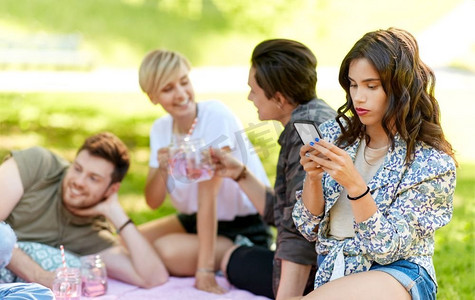 妇女野餐摄影照片_休闲，技术和人的概念—妇女使用智能手机在野餐与朋友在夏天公园。妇女使用智能手机在野餐与朋友