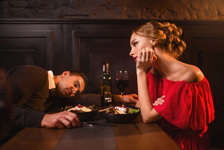 喝醉的男人睡在桌子上反对在餐厅的红色衣服的女人。夫妇有一个破坏的夜晚。喝醉的男人睡在桌子对女人