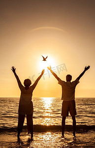 一对老年男女在荒凉的热带海滩上手牵手庆祝日落或日出，一只鸟从太阳旁边飞过。