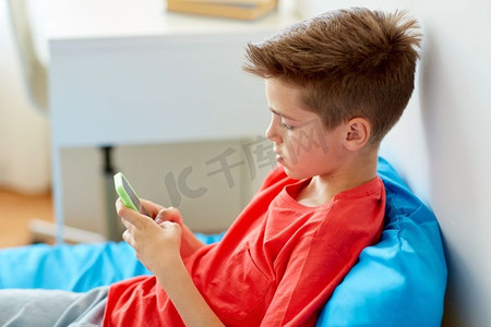 儿童智能摄影照片_童年、网络欺凌和交流概念--家里躺在床上的智能手机男孩。拿着智能手机的男孩躺在家里的床上