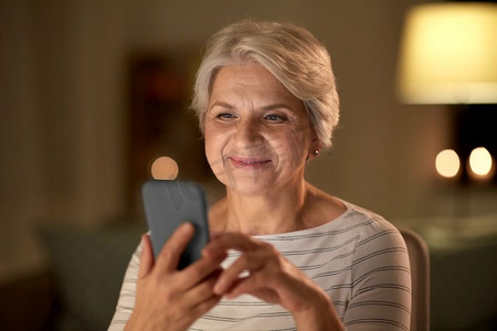 科技、通讯和人的概念快乐的老年女性晚上在家拿着智能手机。家里有智能手机的快乐老年女性