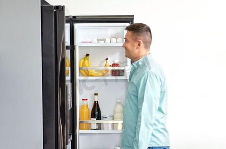 饮食和饮食概念—微笑的中年男子寻找食物在冰箱在厨房。男人在厨房的冰箱里寻找食物