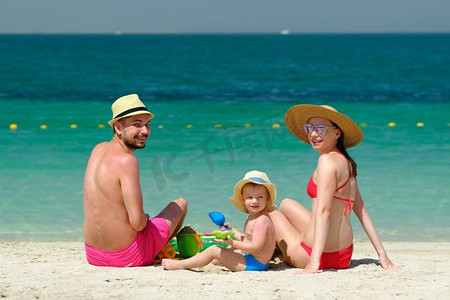 海滩上的家庭使用与海滩玩具的两岁蹒跚学步的男孩与母亲和父亲在海滩