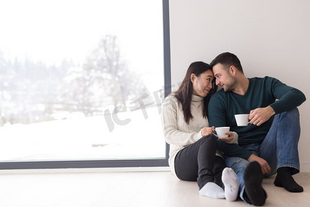 在寒冷的冬日，幸福的年轻多民族夫妇在家中靠窗喝着早晨的咖啡