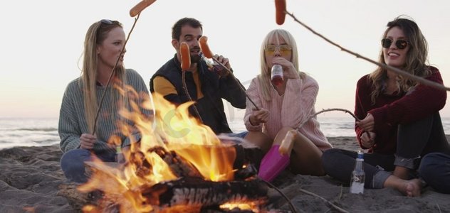 深夜烧烤摄影照片_一群年轻的朋友深夜坐在火堆旁，烤着香肠，喝着啤酒，聊天，玩得很开心
