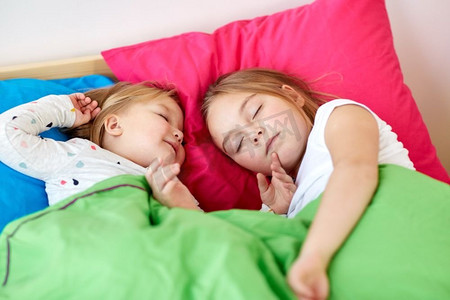 睡觉小女孩摄影照片_童年，睡觉时间和家庭概念-快乐的小女孩或姐妹睡在家里的床上。快乐的小女孩们睡在家里的床上