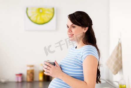 怀孕，人们和期待的概念--快乐的孕妇在家中的厨房里喝茶。快乐的孕妇在家中厨房里拿着杯子