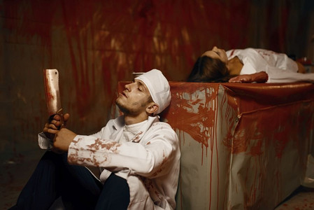 疯子拿着切肉刀坐在女病人旁边,在精神病院地下室做实验,背景是血腥的