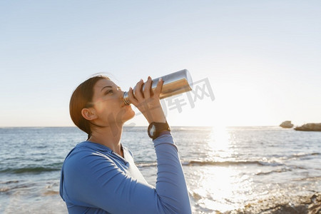 出水素果摄影照片_一名妇女在海滩上喝水。年轻女子在海滩上运动后喝水