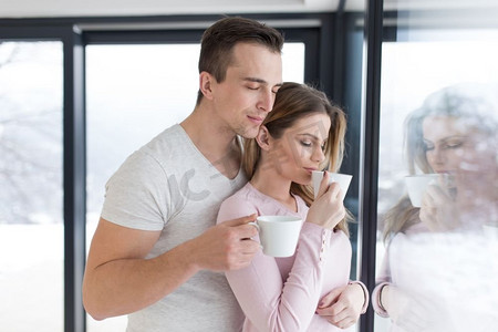 冬日浪漫摄影照片_在寒冷的冬日里，浪漫幸福的年轻夫妇在家中靠窗喝着早晨的咖啡