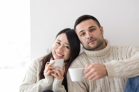 室内寒冷摄影照片_在寒冷的冬日，幸福的年轻多民族夫妇在家中靠窗喝着早晨的咖啡