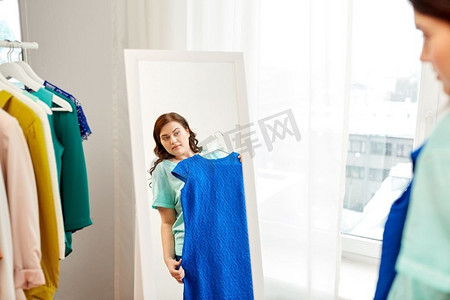 服装时尚和人的概念-沉思的大码女人在家里的镜子里穿蓝色连衣裙。忧郁的大码女人对着镜子穿着蓝色连衣裙
