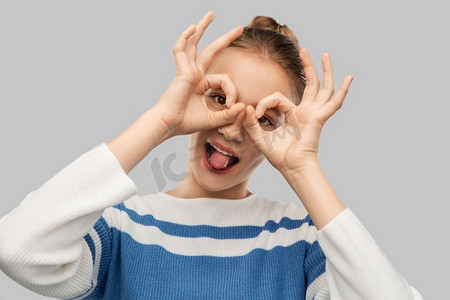 人的概念—滑稽的少女看通过手指眼镜在灰色背景。有趣的十几岁的女孩看通过手指眼镜