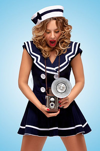 一张复古照片，一位时尚的水手女孩惊讶地看着一台老式照相相机，蓝色背景上有灯泡闪光灯。