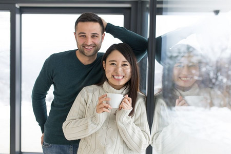 冬日浪漫摄影照片_在寒冷的冬日，幸福的年轻多民族夫妇在家中靠窗喝着早晨的咖啡