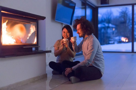 年轻浪漫的多民族情侣坐在家里壁炉前的地板上，在寒冷的冬夜里聊天喝茶