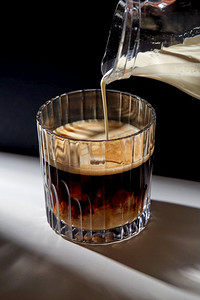 咖啡因和饮料概念--用水壶将奶油或牛奶倒到桌子上的一杯咖啡中。手拿着罐子把奶油倒到一杯咖啡里