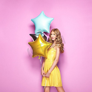 粉色背景活动摄影照片_美丽的年轻女子，粉色背景上有彩色气球。生日和派对。节日上穿着黄色衣服的女孩。情人节