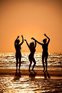 朋友聚会剪影摄影照片_日落时分，三个穿着比基尼的漂亮年轻女子在海滩上跳舞，全都在剪影中