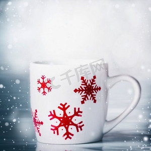 蓝色雪花图摄影照片_白色马克杯，红色雪花，蓝色雪花背景，前景。寒假快乐卡片排版