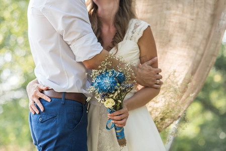 抱花的摄影照片_新娘拥抱新郎，手持印有蓝色花朵的新娘花束，背景是森林