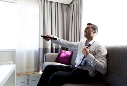 人和休息的概念-在酒店房间里，电视遥控器脱下领带的快乐商人。快乐的商人在酒店房间里拿着电视遥控器