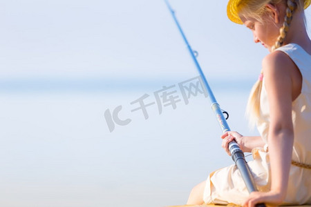女孩穿着裙子，戴着一顶带钓鱼竿的帽子。穿着连衣裙戴着帽子的女孩在码头钓鱼