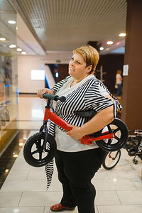 胖女人拿着小自行车在商场。超重女性与孩子自行车，在超市购物，肥胖问题