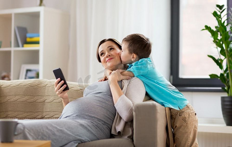孕妈摄影照片_家庭、情感与孕期观念--小儿子在家用智能手机亲吻快乐的孕妈。儿子在家中亲吻幸福的怀孕母亲