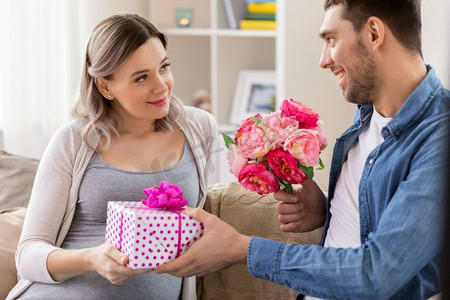 节日、怀孕和问候的概念-快乐的男人在家给怀孕的女人送花。男子在家中向孕妇献花