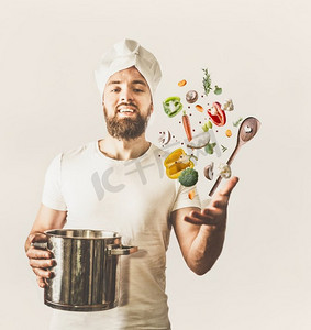 厨师胡子摄影照片_年轻厨师人与帽子和胡子举行平底锅和勺子与飞行蔬菜在白色背景。  健康的烹饪理念