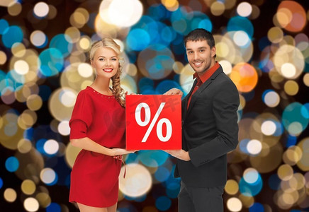 购物，销售和人们的概念快乐的夫妇与圣诞彩灯背景打折标志。在圣诞彩灯上挂着打折标志的夫妇