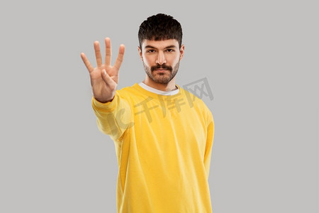 黄色风格摄影照片_伯爵和人的概念—年轻人在黄色运动衫显示四个手指在灰色背景。身穿黄色运动衫的男子露出四根手指