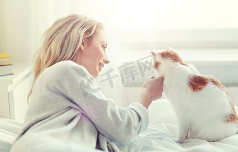 宠物，早晨，舒适，休息和人概念-有猫的愉快的少妇在家床上。快乐的年轻女子与猫在床上在家