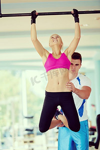 支持配合摄影照片_在室内健身房的单杠上举重时，教练在支撑年轻女子。教练在健身房的单杠上举重时支持年轻女性