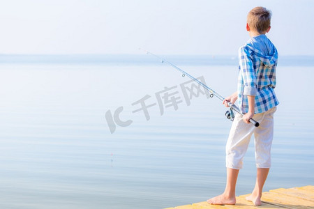 手里拿鱼摄影照片_穿蓝衬衫的男孩站在馅饼上。穿着蓝色衬衫的男孩站在海边的码头上，手里拿着一根钓鱼竿