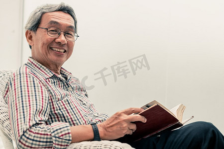 愉快的高级亚洲人读书在椅子上的书在家里的客厅。退休生活方式与老龄化社会观念。