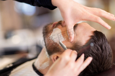 剃胡子的男人摄影照片_美容和人的概念-理发店里留着直剃刀胡子的男人和理发师。留着直剃须刀的男人和理发师